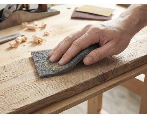 Hand die een houten plank bewerkt met schuurvlies op een lichte werkbank met een houtschaaf en houtkrullen.