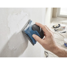 Een hand schuurt een geplamuurde muur glad met een handschuurmachine en schuurvlies.