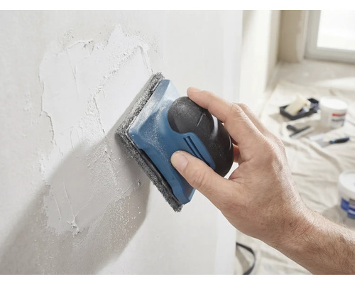 Een hand schuurt een geplamuurde muur glad met een handschuurmachine en schuurvlies.