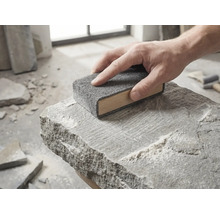 Een hand beweegt een houten schuurblok met grijs schuurvlies over het oppervlak van een ruwe natuursteen.