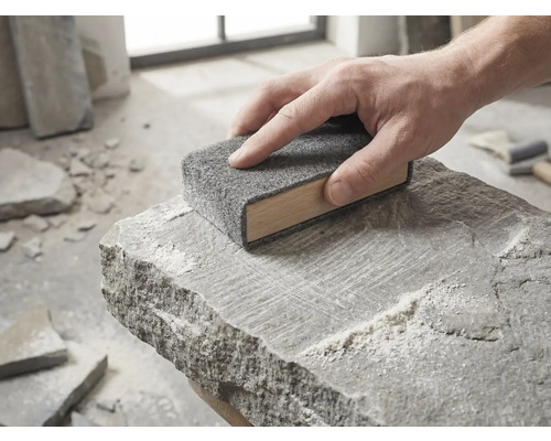 Een hand beweegt een houten schuurblok met grijs schuurvlies over het oppervlak van een ruwe natuursteen.