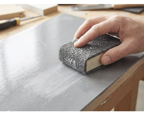 Een hand schuurt een gelakt houten oppervlak met een schuurvlies dat om een schuurblok is gewikkeld.