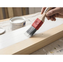 Een kwast met houten steel en rode huls brengt witte verf aan op een houten balk. Op de achtergrond staat een open verfblik.
