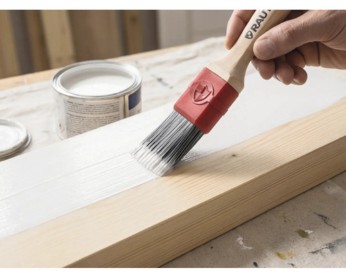 Een kwast met houten steel en rode huls brengt witte verf aan op een houten balk. Op de achtergrond staat een open verfblik.