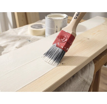 Een kwast met een houten steel brengt witte verf aan op een houten plank, op de achtergrond zijn een verfblik en schilderstape te zien.