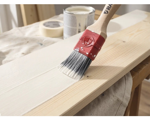 Een kwast met een houten steel brengt witte verf aan op een houten plank, op de achtergrond zijn een verfblik en schilderstape te zien.