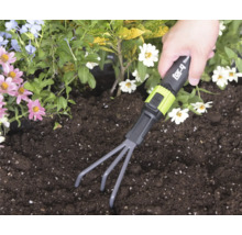 Een hand gebruikt een for_q handhark in donkere aarde in een tuinbed met bloemen.