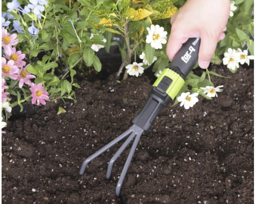 Een hand gebruikt een for_q handhark in donkere aarde in een tuinbed met bloemen.