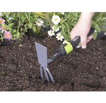 Hand hanteert een for_q handhak met tanden en blad in donkere aarde naast bloemen om de grond los te maken.