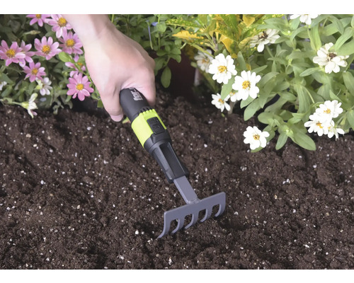 Handhark met zwart en groen handvat en for_q Logo wordt gebruikt in donkere potgrond tussen roze en witte bloemen.