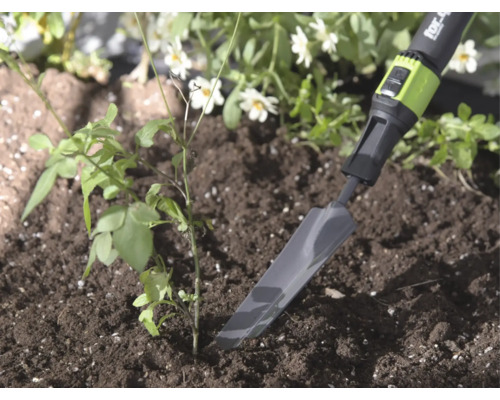 Smalle metalen verplantschop van for_q in de aarde naast een kleine plant, lichte en natuurlijke tuinomgeving.