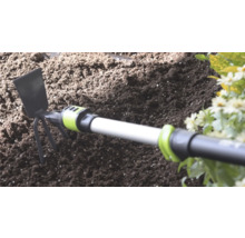 Handhak met metalen blad en drie tanden in donkere tuingrond naast bloemen.