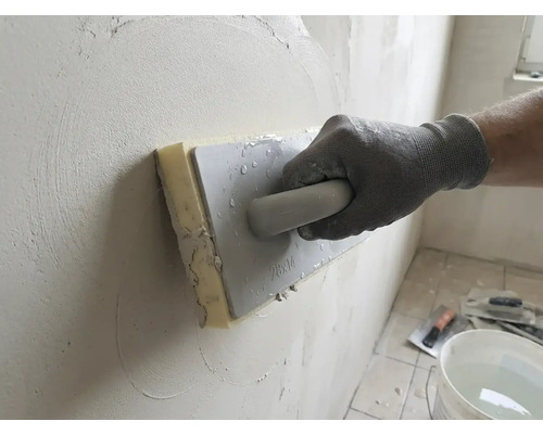 Hand met werkhandschoen gebruikt een sponsspaan voor het gladstrijken van stucwerk op een muur.