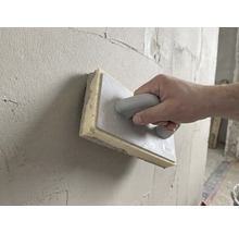 Hand gebruikt een sponsspaan van 28 bij 14 centimeter om vers pleisterwerk op een muur glad te strijken.