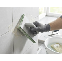 Een persoon voegt wandtegels met een voegbord. Op de achtergrond staan een emmer met water en een spons en een plakspaan.