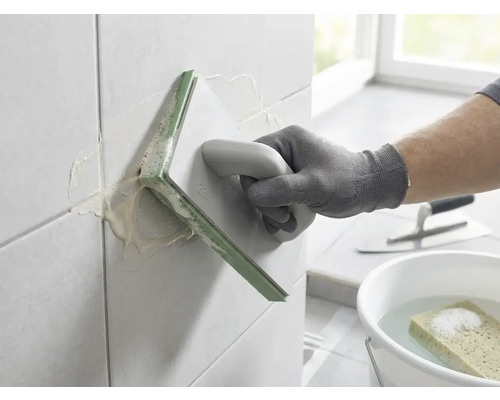 Een persoon voegt wandtegels met een voegbord. Op de achtergrond staan een emmer met water en een spons en een plakspaan.