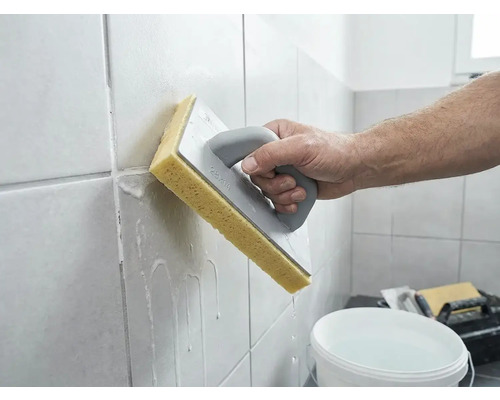 Persoon reinigt vers gevoegde wandtegels met een sponsbord en water.