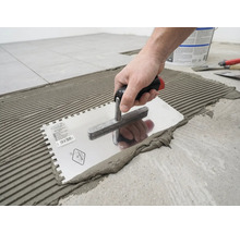 Een hand brengt tegellijm aan op de vloer met een getande lijmkam in een geribbeld patroon.