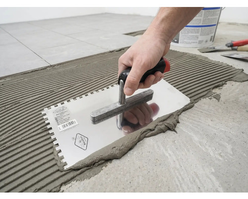 Een hand brengt tegellijm aan op de vloer met een getande lijmkam in een geribbeld patroon.