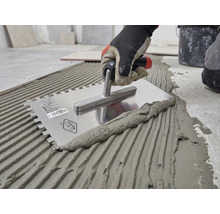 Hand met werkhandschoen brengt tegellijm aan op de vloer met een roestvrijstalen lijmkam.