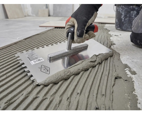 Hand met werkhandschoen brengt tegellijm aan op de vloer met een roestvrijstalen lijmkam.