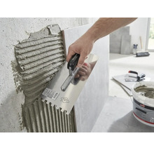 Een hand brengt tegellijm aan op een muur met een lijmkam.