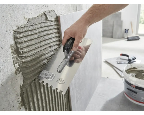 Een hand brengt tegellijm aan op een muur met een lijmkam.