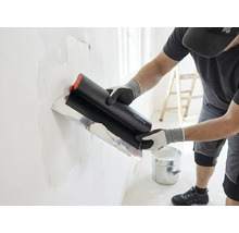 Vakman brengt met een breed plamuurmes plamuur aan op een muur, in een lichte ruimte staan een emmer en een ladder.