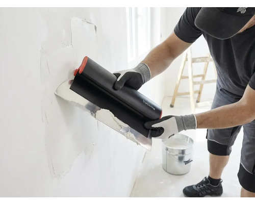 Vakman brengt met een breed plamuurmes plamuur aan op een muur, in een lichte ruimte staan een emmer en een ladder.