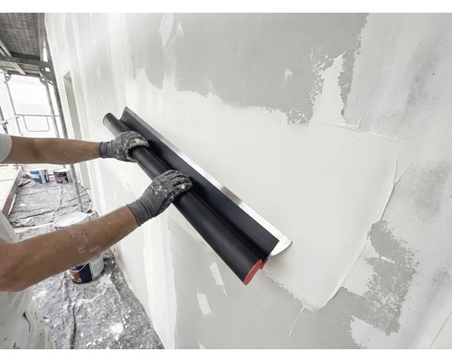 Persoon gebruikt een brede pleisterspaan met roestvrijstalen blad om stucwerk op een muur glad te strijken tijdens renovatie.