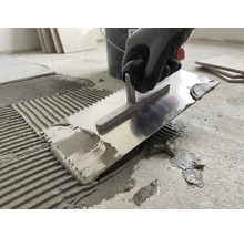 Een persoon brengt met een metalen lijmkam tegellijm aan op een betonnen vloer.