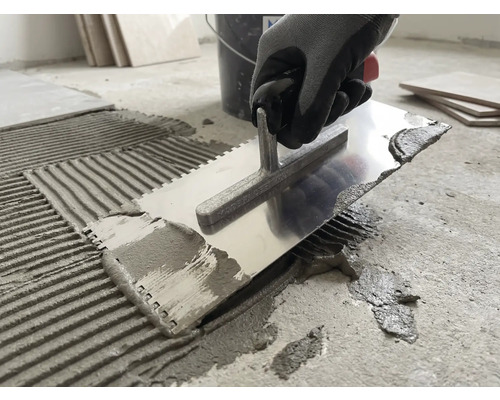 Een persoon brengt met een metalen lijmkam tegellijm aan op een betonnen vloer.