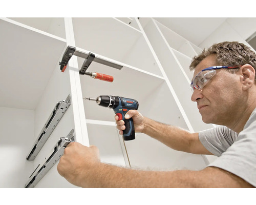 Een man met een veiligheidsbril monteert ladegeleiders in een witte kast met een Bosch accuboorschroevendraaier en een lijmtang.