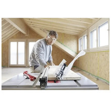 Een man met veiligheidsbril zaagt een houten plank met een tafelcirkelzaag in een lichte ruimte met houten balken en OSB-wanden.