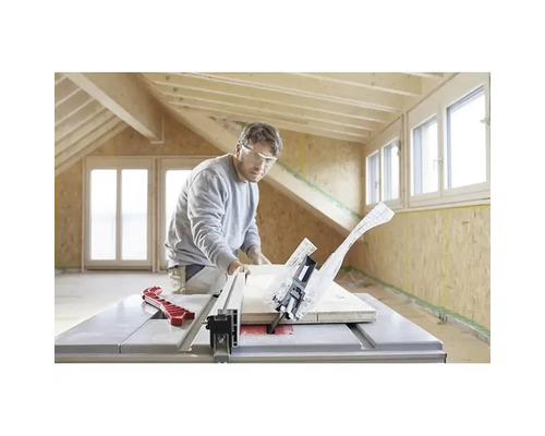 Een man met veiligheidsbril zaagt een houten plank met een tafelcirkelzaag in een lichte ruimte met houten balken en OSB-wanden.