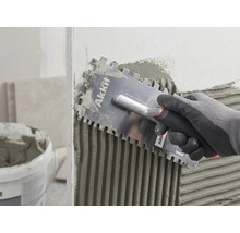 Een hand met handschoen brengt met een Akkit lijmkam tegellijm gelijkmatig aan op een muur.