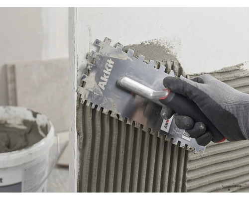 Een hand met handschoen brengt met een Akkit lijmkam tegellijm gelijkmatig aan op een muur.