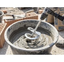 Een menger op een boormachine mengt grijze mortel in een bouwemmer op een bouwplaats met bakstenen en troffels.