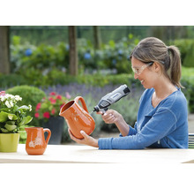 Vrouw met veiligheidsbril graveert een kan van terracotta met een Dremel multifunctioneel gereedschap in een lichte tuin.