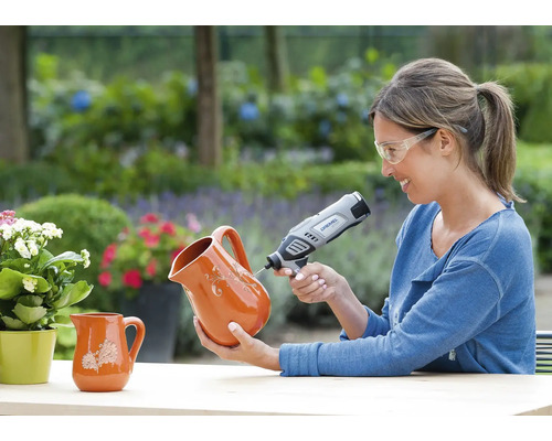 Vrouw met veiligheidsbril graveert een kan van terracotta met een Dremel multifunctioneel gereedschap in een lichte tuin.