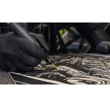 Hand in zwarte handschoen graveert een fijn patroon op een donker oppervlak met een multifunctioneel gereedschap.