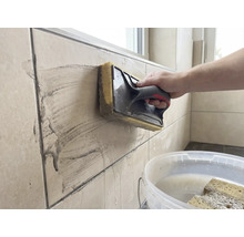 Een persoon reinigt beige wandtegels met een sponsspaan. Op de voorgrond staat een witte emmer met water.