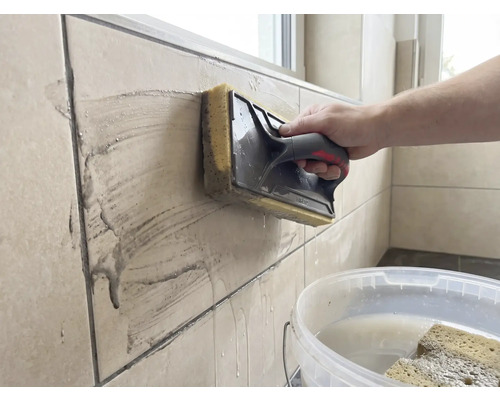 Een persoon reinigt beige wandtegels met een sponsspaan. Op de voorgrond staat een witte emmer met water.