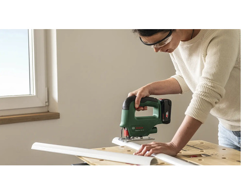 Persoon met veiligheidsbril gebruikt een Bosch accu decoupeerzaag voor het zagen van een wit kunststof profiel op een werkbank.