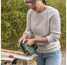 Vrouw met veiligheidsbril zaagt een wit houten profiel met een Bosch draadloze decoupeerzaag EasySaw 18 Volt op een werkbank in een lichte tuin.