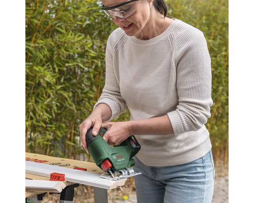 Vrouw met veiligheidsbril zaagt een wit houten profiel met een Bosch draadloze decoupeerzaag EasySaw 18 Volt op een werkbank in een lichte tuin.
