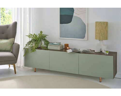 Lichte, modern ingerichte woonkamer met een dressoir in groen en houtlook, een grijze fauteuil, een kamerplant en een tafellamp.