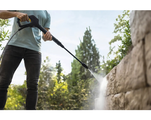 Een persoon reinigt een gestructureerde stenen muur met de spuitlans van een hogedrukreiniger in de tuin.