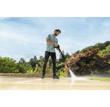 Een man reinigt een licht stenen terras in de tuin met een hogedrukreiniger bij zonnig weer.