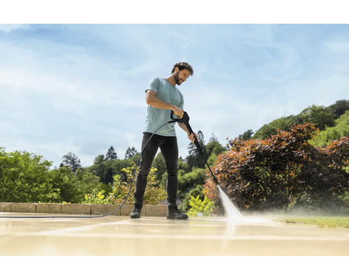 Een man reinigt een licht stenen terras in de tuin met een hogedrukreiniger bij zonnig weer.